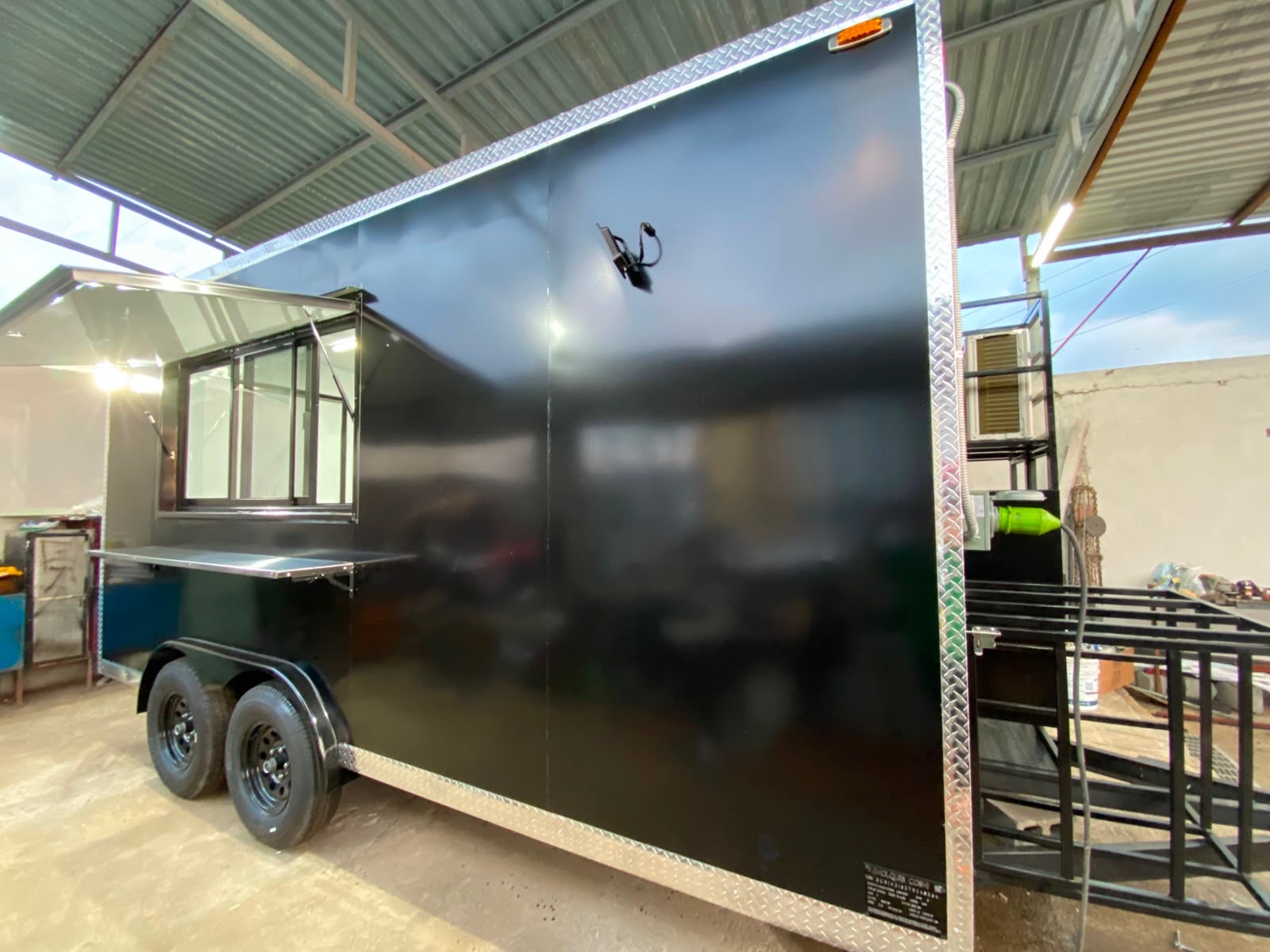Full flank of the food truck in the shop, showing the gloss black body and diamond-plate trim over the tandem axle.