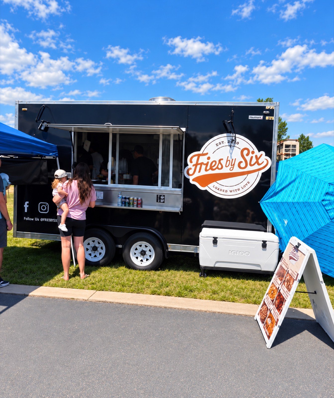 Fries by Six food truck set up on grass at a daytime event, a family ordering at the window under a canopy.