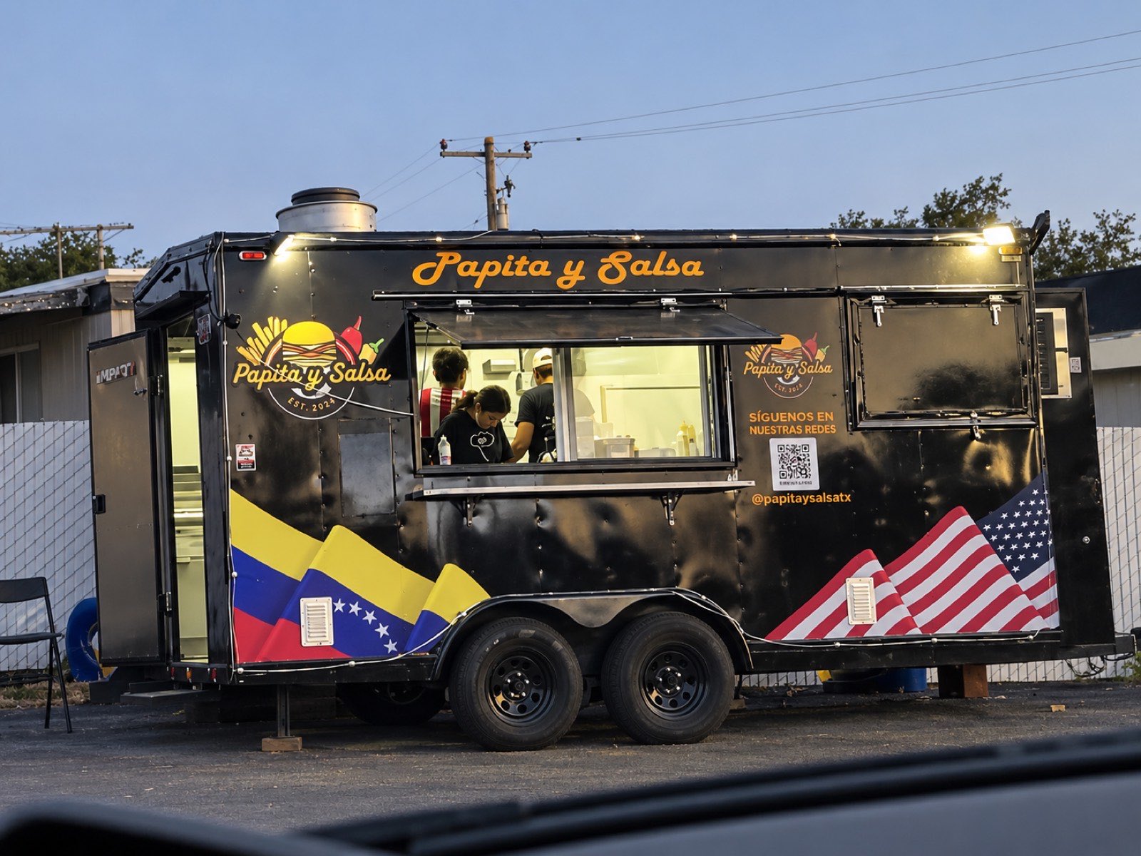 Full flank of the Papita y Salsa food truck at dusk with Venezuelan and US flag livery and the crew working the line.